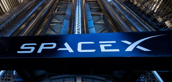 The image shows the SpaceX logo prominently displayed on a large, metallic structure, with an upward perspective that highlights the industrial and futuristic design of the facility. The setting suggests advanced aerospace technology and innovation, characteristic of SpaceX's mission in space exploration.