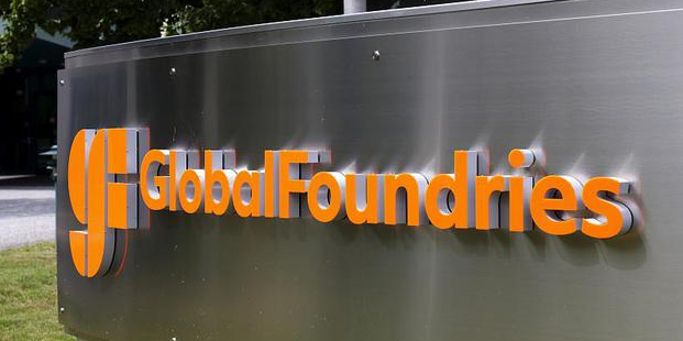 A brushed metal sign featuring the GlobalFoundries logo and name in bold orange letters, mounted outdoors with trees visible in the background.