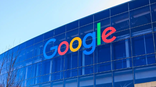 The exterior of a modern Google office building with the colourful Google logo displayed prominently on the glass facade under a clear blue sky.