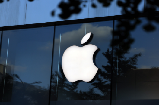 A large, illuminated Apple logo displayed on the glass facade of an Apple Store, with reflections of trees and the sky visible in the background.